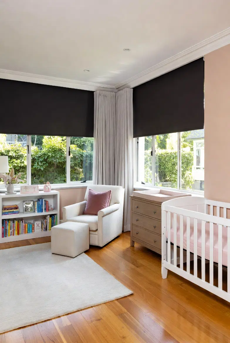 Blockout roller blinds in a nursery with cot, armchair, and garden views through half-raised blinds