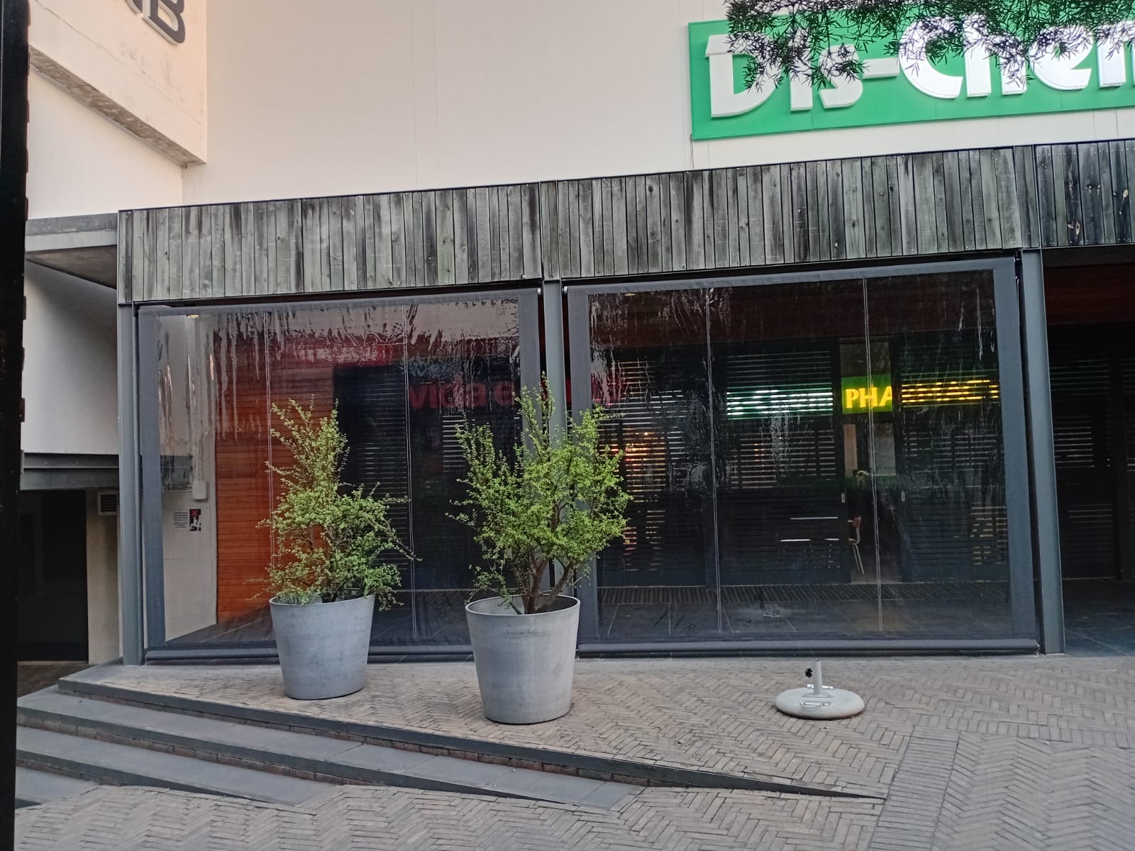 Exterior view of clear PVC blinds at Vida e Cafe Knysna Mall with Dis-Chem signage above, fully enclosing the outdoor seating terrace