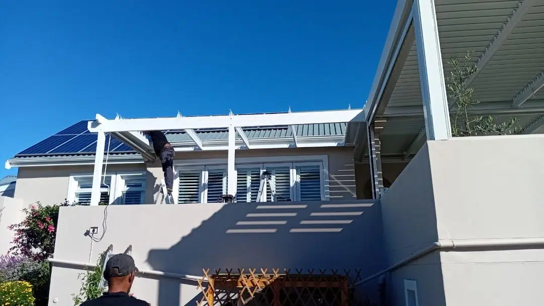 Custom Blinds installation team fitting an aluminium louvre awning frame to a Knysna home. Open blades visible against blue sky during installation. Garden Route, May 2026.
