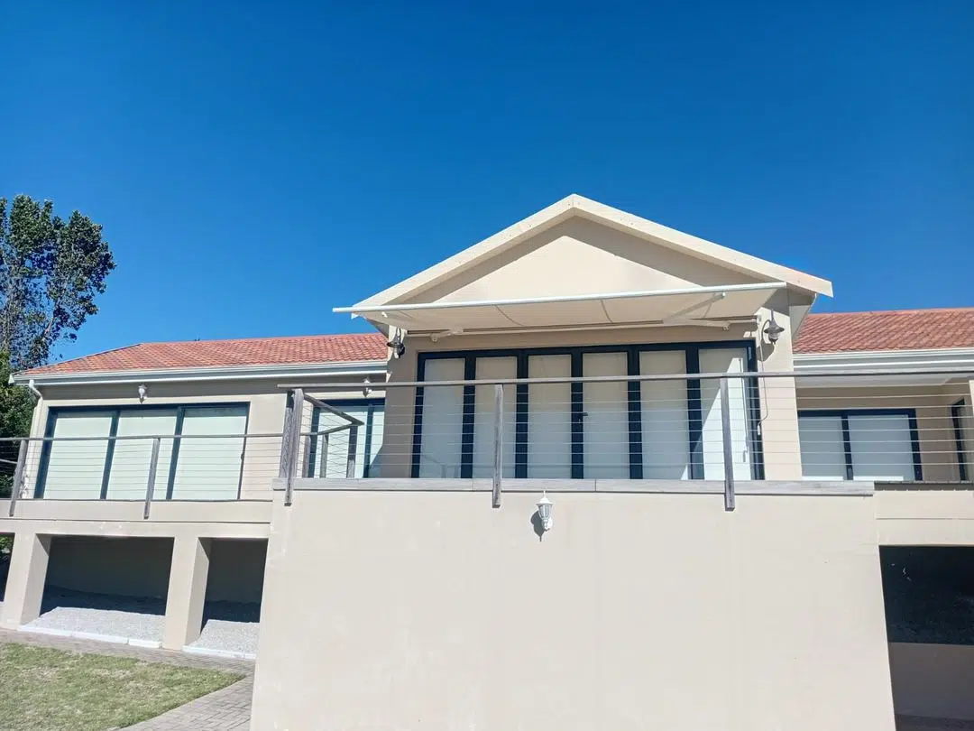 Cream cassette retractable folding-arm awning extended over a first-floor balcony of a Plettenberg Bay home, mounted under a gable roof. Custom Blinds Garden Route installation, May 2026.
