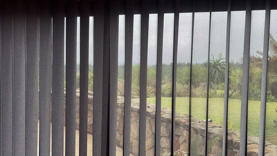 Vertishades sheer voile vertical blinds across a large window in a Garden Route home, filtering a view of a stone garden wall, lawn and aloes