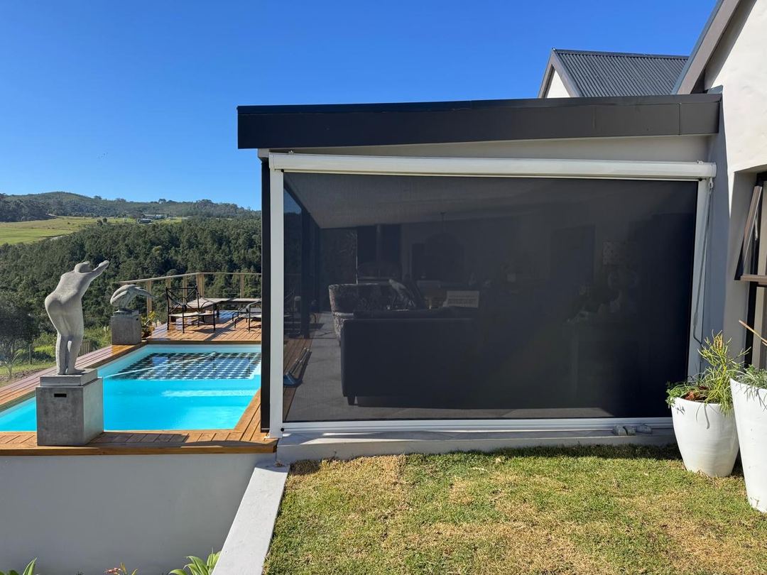 Dark screen mesh sidetrack outdoor blind closing a covered patio beside a pool deck, by Custom Blinds Shutters & Awnings, Knysna